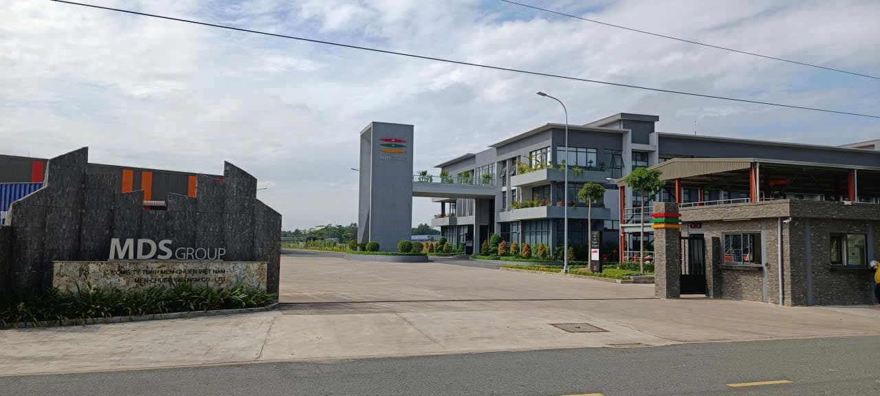 Cổng nhà máy Men-Chuen Việt Nam (MDS Group) trong Khu công nghiệp Nhơn Trạch 6: khối văn phòng nhiều tầng có cây xanh trên ban công, bảng hiệu đá bên trái cổng